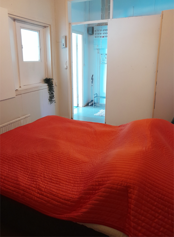 A bed with a red blanket on it, the room's door is open to the hallway